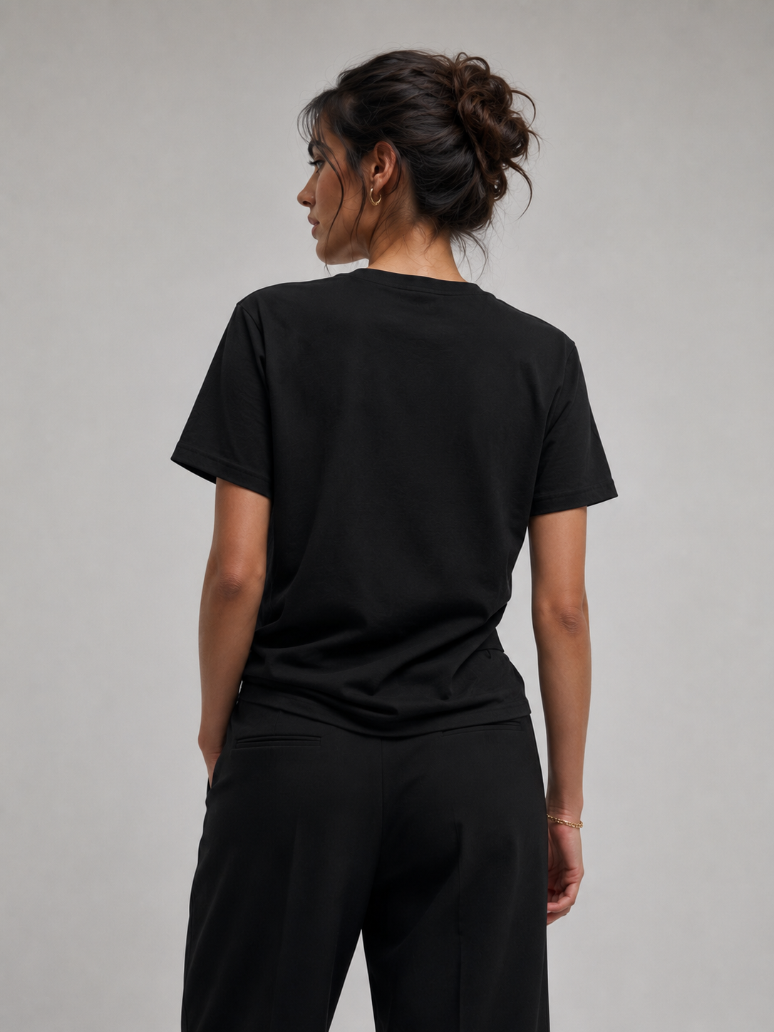 A woman shown from the back wearing a black short-sleeve crewneck t-shirt and black pants against a plain light gray studio background.
