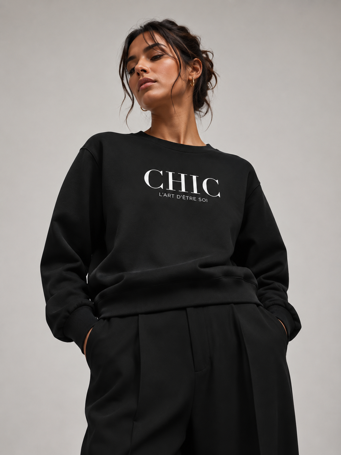 A woman wearing a black crewneck sweatshirt with 'CHIC L'ART D'UNE SOI' printed in white on the chest, paired with black trousers, against a neutral gray background.