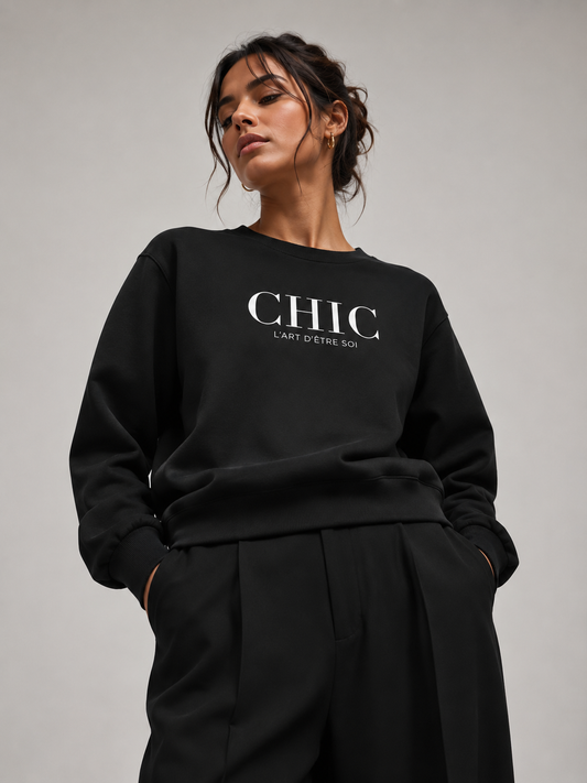 A woman wearing a black crewneck sweatshirt with 'CHIC L'ART D'UNE SOI' printed in white on the chest, paired with black trousers, against a neutral gray background.