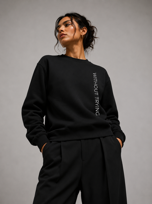 A woman shown front wearing a black crewneck sweatshirt with a graphic and the phrase “without trying” on the back, paired with black pants in a minimalist light gray studio.