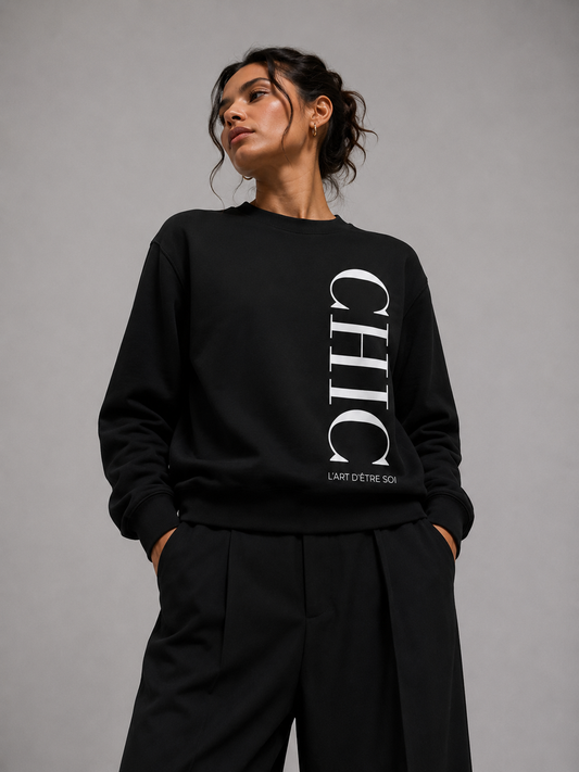 A woman wearing a black crewneck sweatshirt with a large white vertical 'CHIC L'art D'etre soi' graphic and matching black wide-leg trousers, standing against a neutral gray studio background.