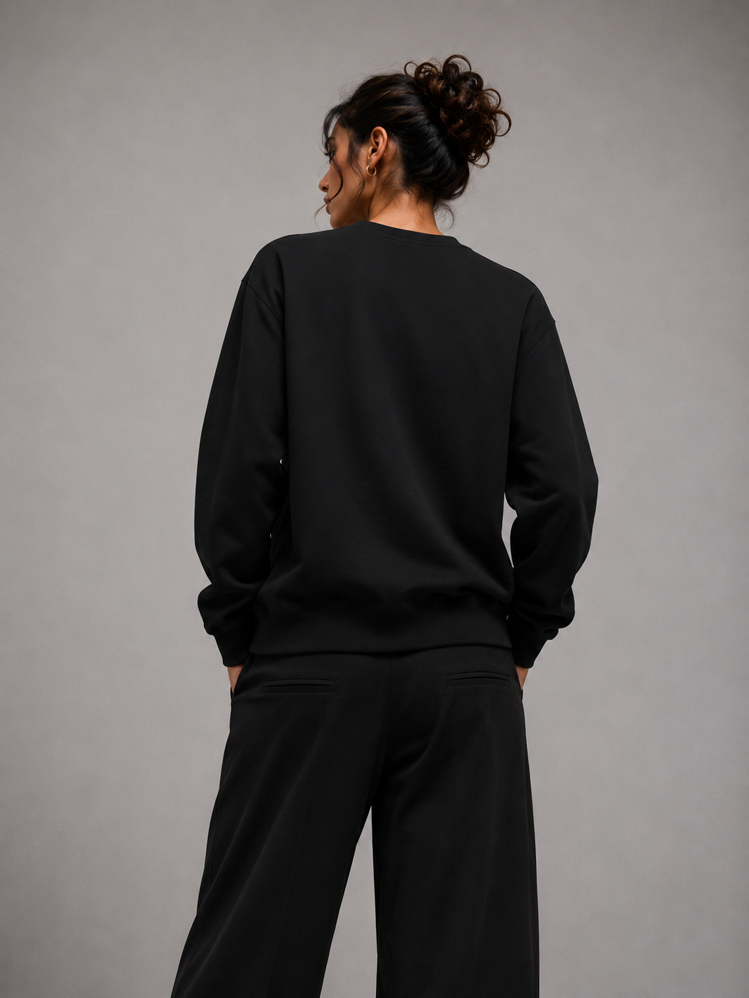 A person shown from the back wearing a black crewneck sweatshirt and black wide-leg pants against a plain gray studio background.