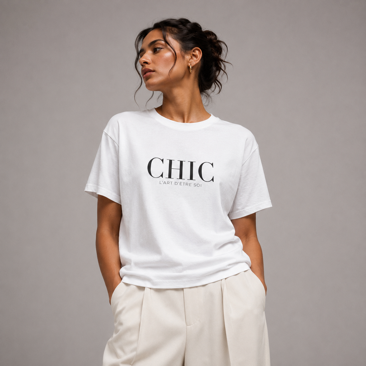 A woman wearing a white crewneck t-shirt with 'CHIC' printed in black across the chest, paired with cream-colored trousers, set against a solid gray studio background.