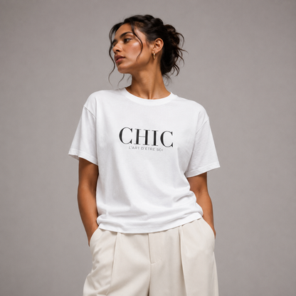 A woman wearing a white crewneck t-shirt with 'CHIC' printed in black across the chest, paired with cream-colored trousers, set against a solid gray studio background.
