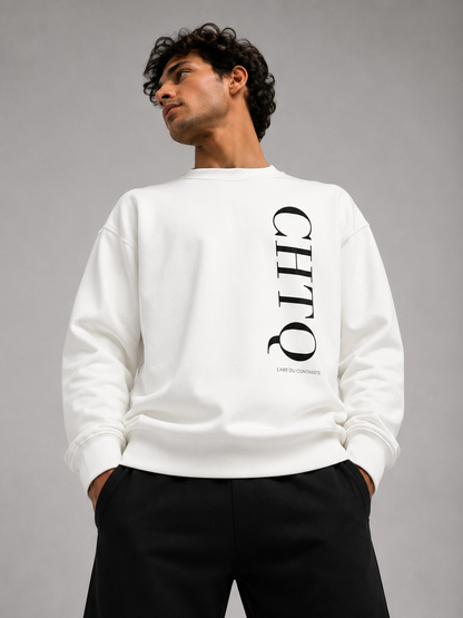 A man wearing a white crewneck sweatshirt with a large vertical black CHTQ graphic on the front, paired with black pants against a neutral gray studio background.