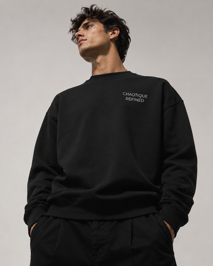A man wearing a black crewneck sweatshirt with 'CHAOTIQUE REFINED' printed in white on the left chest, paired with black pants, against a neutral gray background.