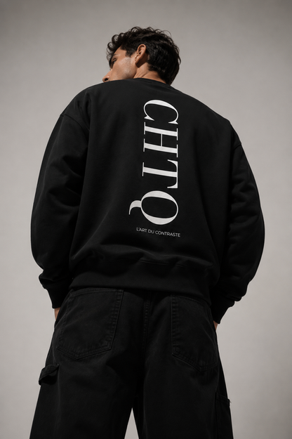 Back view of a man wearing a black crewneck sweatshirt featuring a large white vertical "CHTQ" graphic and "L'Art du Contraste" on the back, photographed in a minimalist gray studio.
