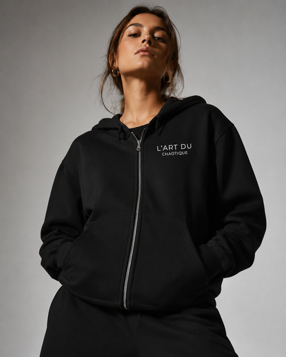 A woman wearing a black zip-up hoodie featuring the white "L'ART DU CHAOTIQUE" graphic on the left chest, photographed against a plain light gray studio background.