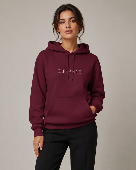 Woman wearing a burgundy ÉLÉGANCE pullover hoodie with white front typography, paired with black trousers in a neutral gray studio setting.
