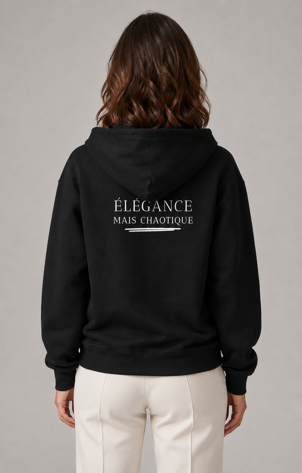 A woman viewed from the back wearing a black pullover hoodie with the French text 'ÉLEGANCE MAIS CHAOTIQUE' printed in white across the upper back, paired with off-white trousers, against a neutral gray studio background.