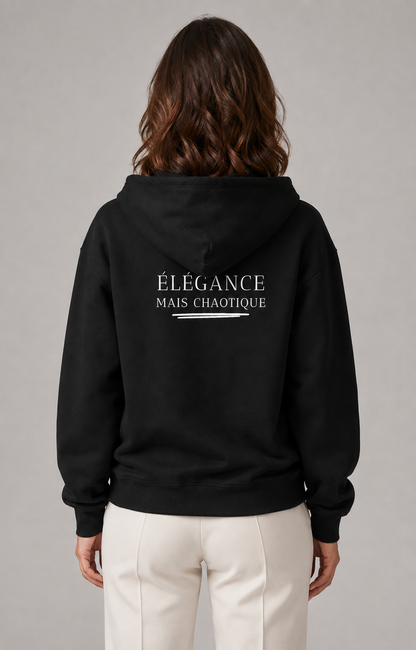A woman viewed from the back wearing a black pullover hoodie with the French text 'ÉLEGANCE MAIS CHAOTIQUE' printed in white across the upper back, paired with off-white trousers, against a neutral gray studio background.