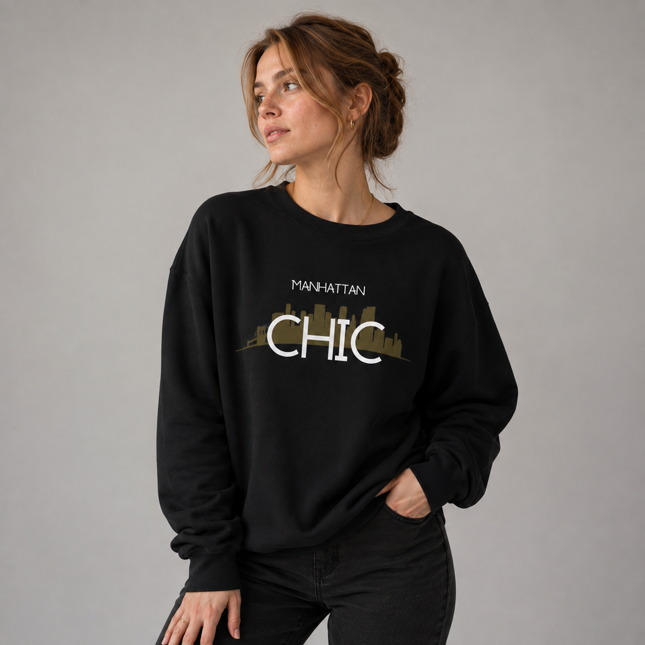 Young woman wearing a black MANHATTAN CHIC crewneck sweatshirt featuring white typography and a muted olive New York skyline graphic, posed against a neutral gray studio background.