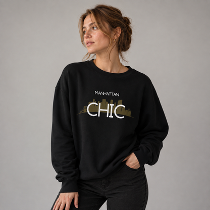 Young woman wearing a black MANHATTAN CHIC crewneck sweatshirt featuring white typography and a muted olive New York skyline graphic, posed against a neutral gray studio background.
