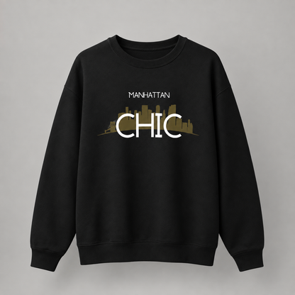 A black crewneck sweatshirt featuring a graphic print on the front with the text 'MANHATTAN CHIC' in white over a muted gold city skyline silhouette, displayed against a neutral gray background.