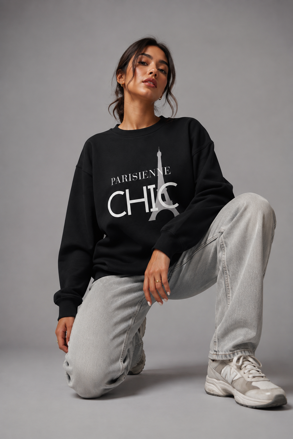 Young woman wearing a black Parisienne CHIC sweatshirt featuring a minimalist Eiffel Tower graphic, kneeling on a gray studio floor with light gray jeans and white sneakers.