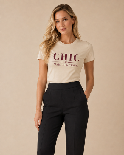 A woman modeling a cream-colored short-sleeve graphic t-shirt with 'CHIC MAIS CHAOTIQUE' printed in burgundy on the front, paired with black trousers, against a solid beige background.