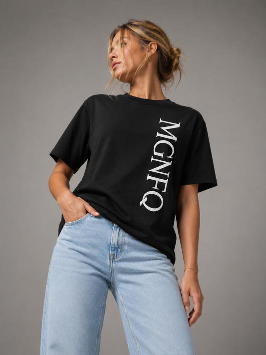 A woman wearing an oversized black T-shirt featuring a vertical white “MGNFQ” print on the side, styled with light blue jeans and photographed against a clean, solid gray studio background.