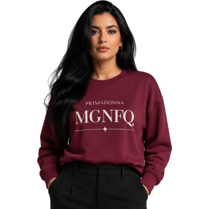 A woman wearing a maroon crewneck sweatshirt with white “PRIMADONNA MGNFQ” text on the chest, paired with black trousers, against a solid gray studio background.