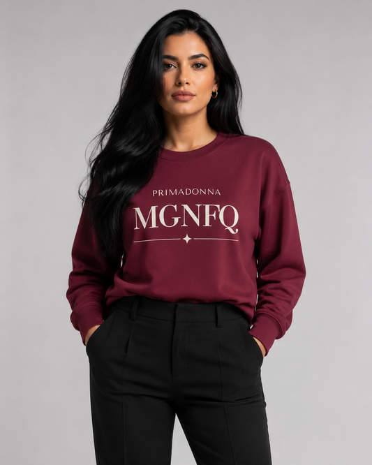 A woman wearing a maroon crewneck sweatshirt with white “PRIMADONNA MGNFQ” text on the chest, paired with black trousers, against a solid gray studio background.
