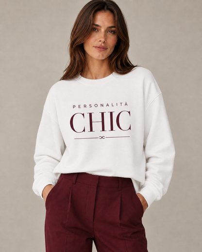 A woman wearing a white crewneck sweatshirt with 'PERSONALITÀ CHIC' printed in dark burgundy on the front, paired with burgundy trousers, set against a neutral gray studio background.