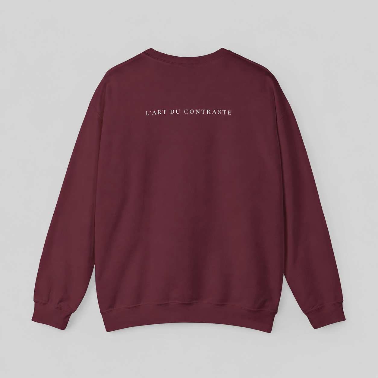 A maroon crewneck sweatshirt shown from the back, featuring the text 'L'ART DU CONTRASTE' printed in small white letters across the upper back, set against a solid light gray background.