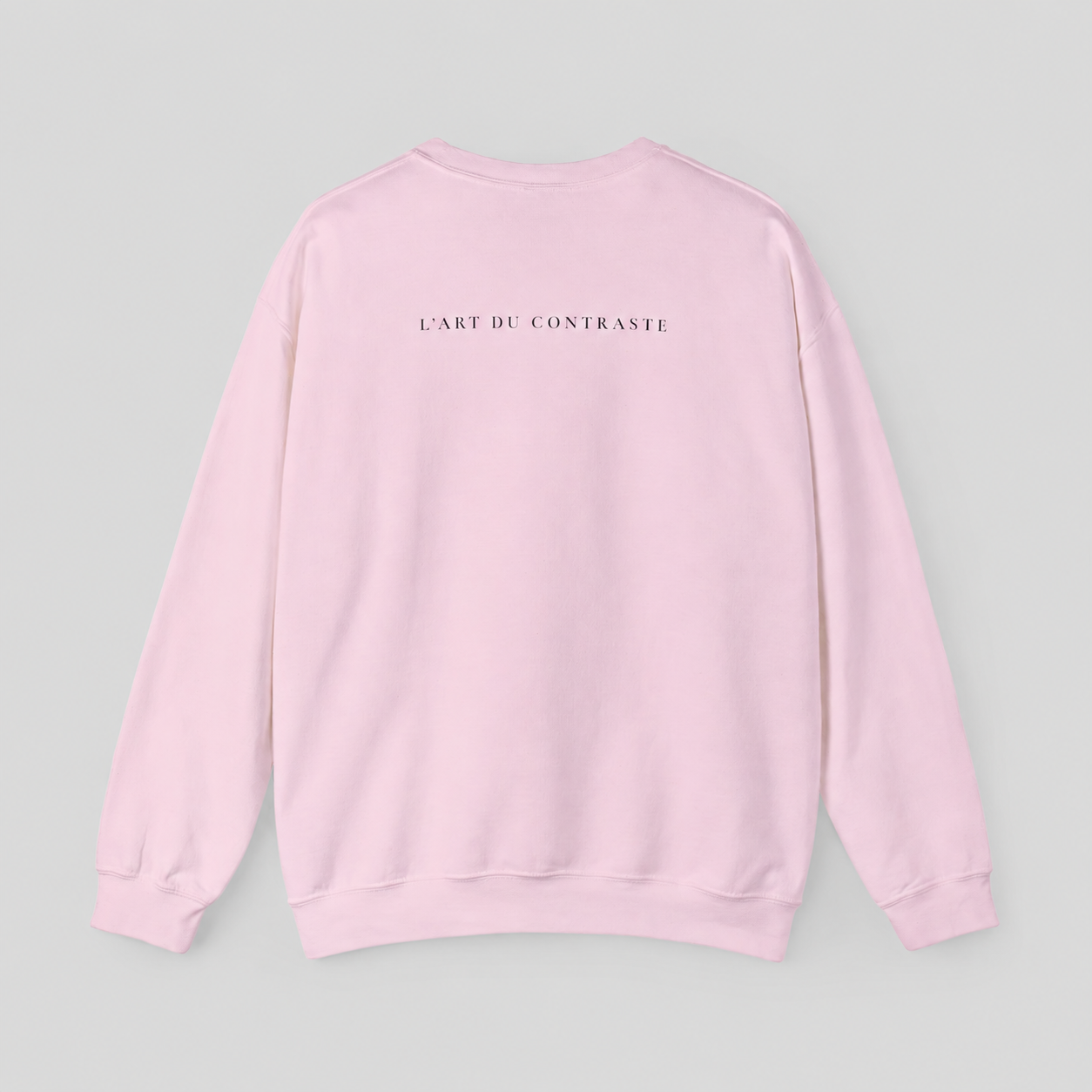 A light pink crewneck sweatshirt shown from the back, featuring the text 'L'ART DU CONTRASTE' printed in small black capital letters across the upper back, set against a solid light gray background.