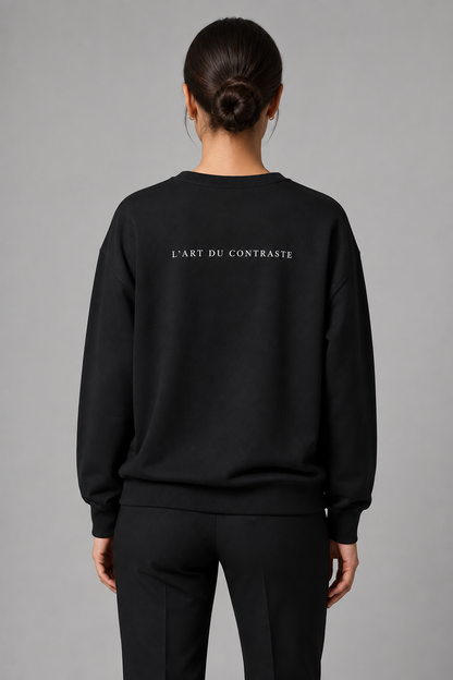 A model shown from the back wearing a black crewneck sweatshirt with the text 'L'ART DU CONTRASTE' printed in white across the upper back, paired with black pants, against a solid gray studio background.
