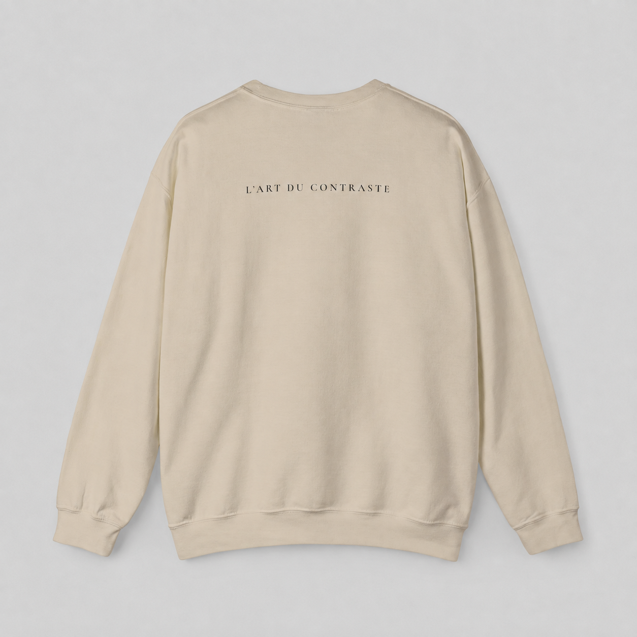 A beige crewneck sweatshirt shown from the back, featuring the text 'L'ART DU CONTRASTE' printed in small black letters across the upper back, set against a neutral light gray background.