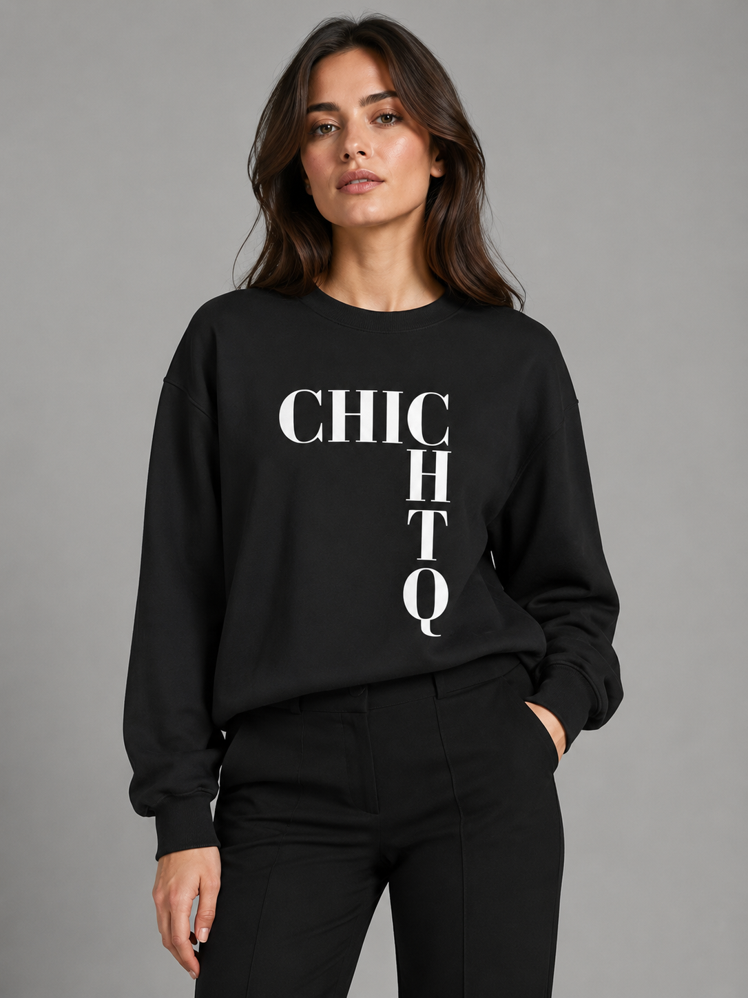 A woman modeling a black crewneck sweatshirt with bold white vertical text reading 'CHIC HQ' on the front, paired with black pants, against a plain gray studio background.