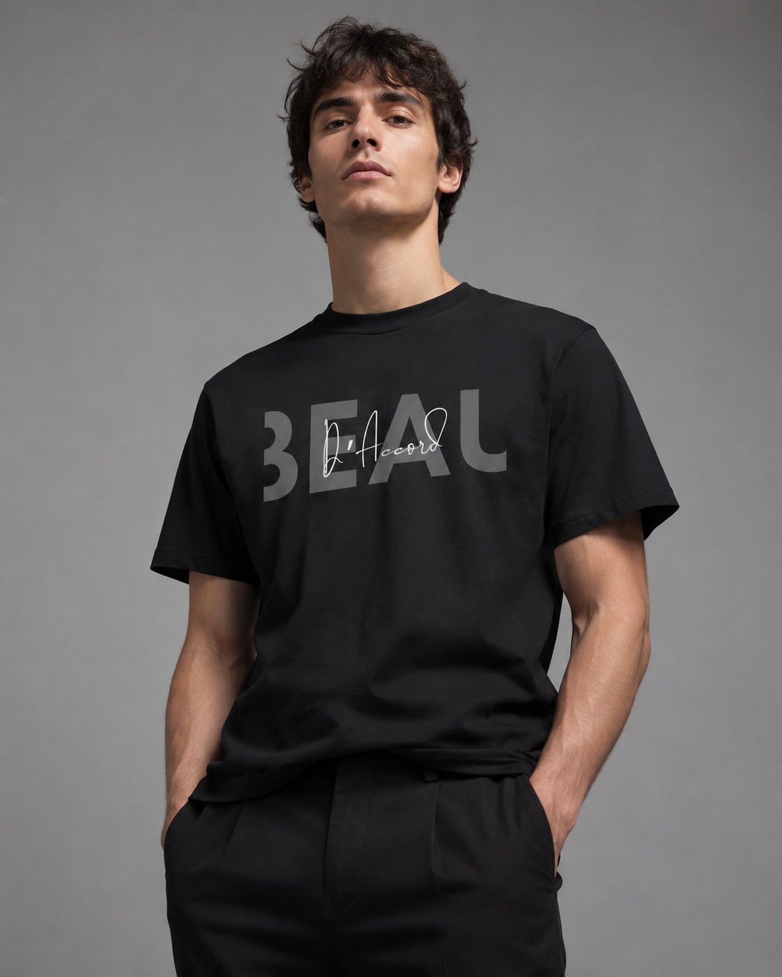 Man wearing a black short-sleeve T-shirt featuring a gray “BEAU” graphic with a white cursive “D’Accord” overlay, posed against a neutral gray studio background.