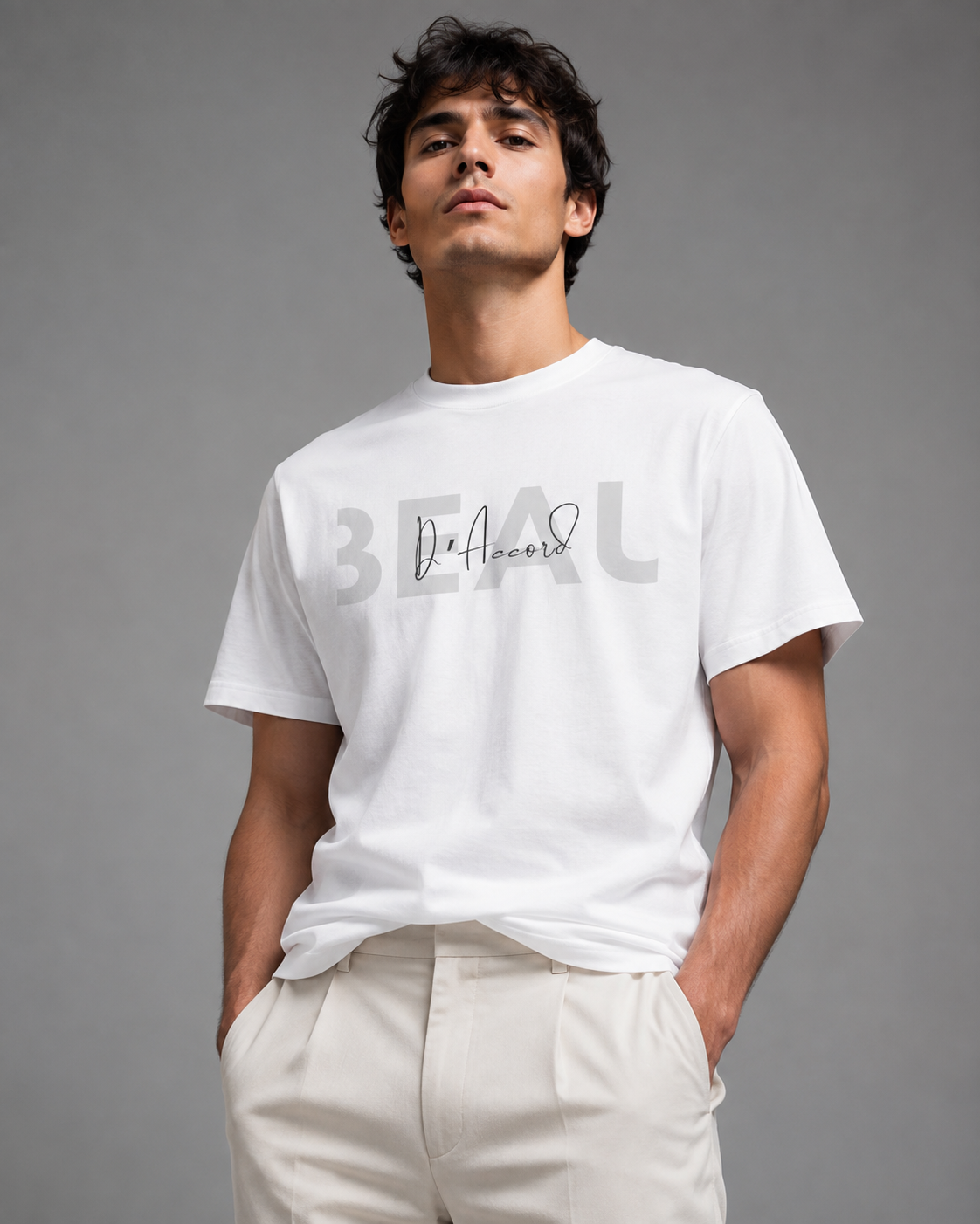 Man wearing a white short-sleeve T-shirt with a gray “BEAU” graphic and black “D’Accord” script, paired with off-white trousers against a gray studio background.