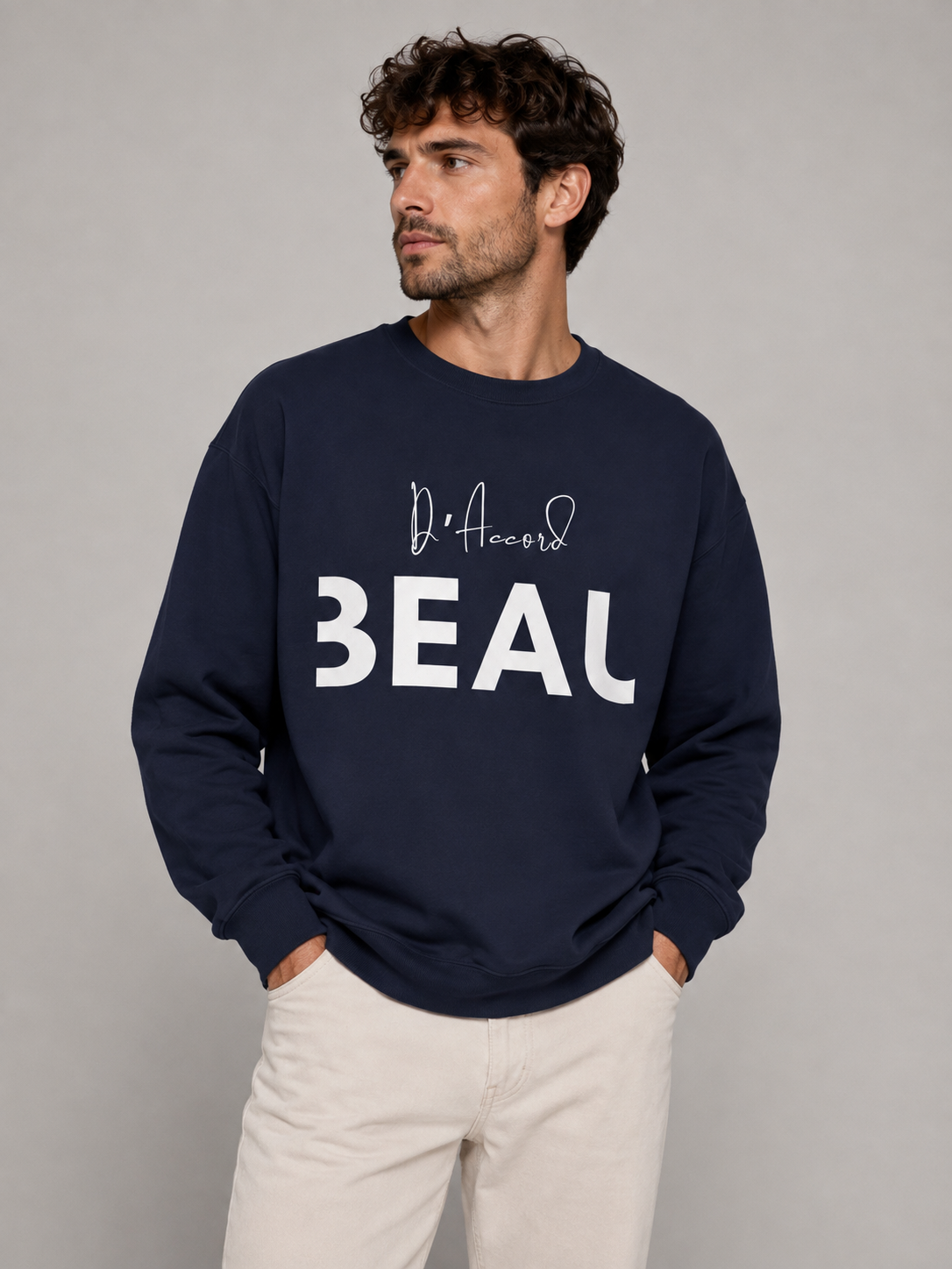 A man wearing a navy blue crewneck sweatshirt featuring a white “D’accord BEAU” graphic print on the front, paired with off-white trousers, against a light gray studio background.