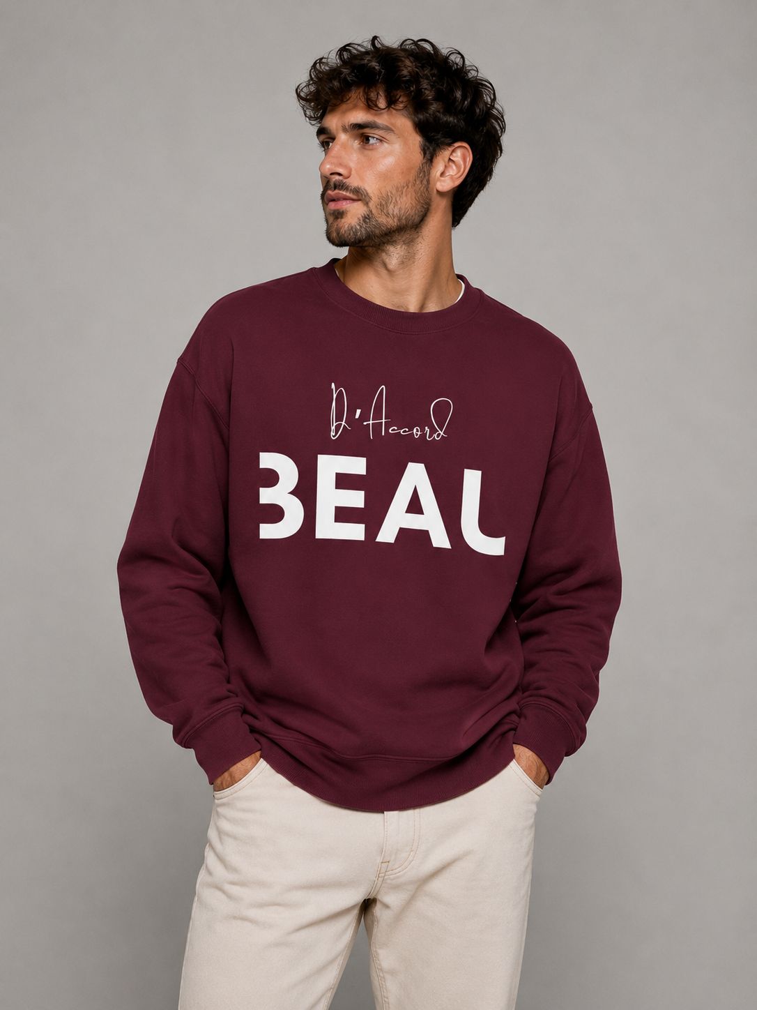 A man wearing a maroon crewneck sweatshirt featuring a black “D’accord” script above bold “BEAU” lettering across the chest, paired with beige trousers, against a light gray studio background.