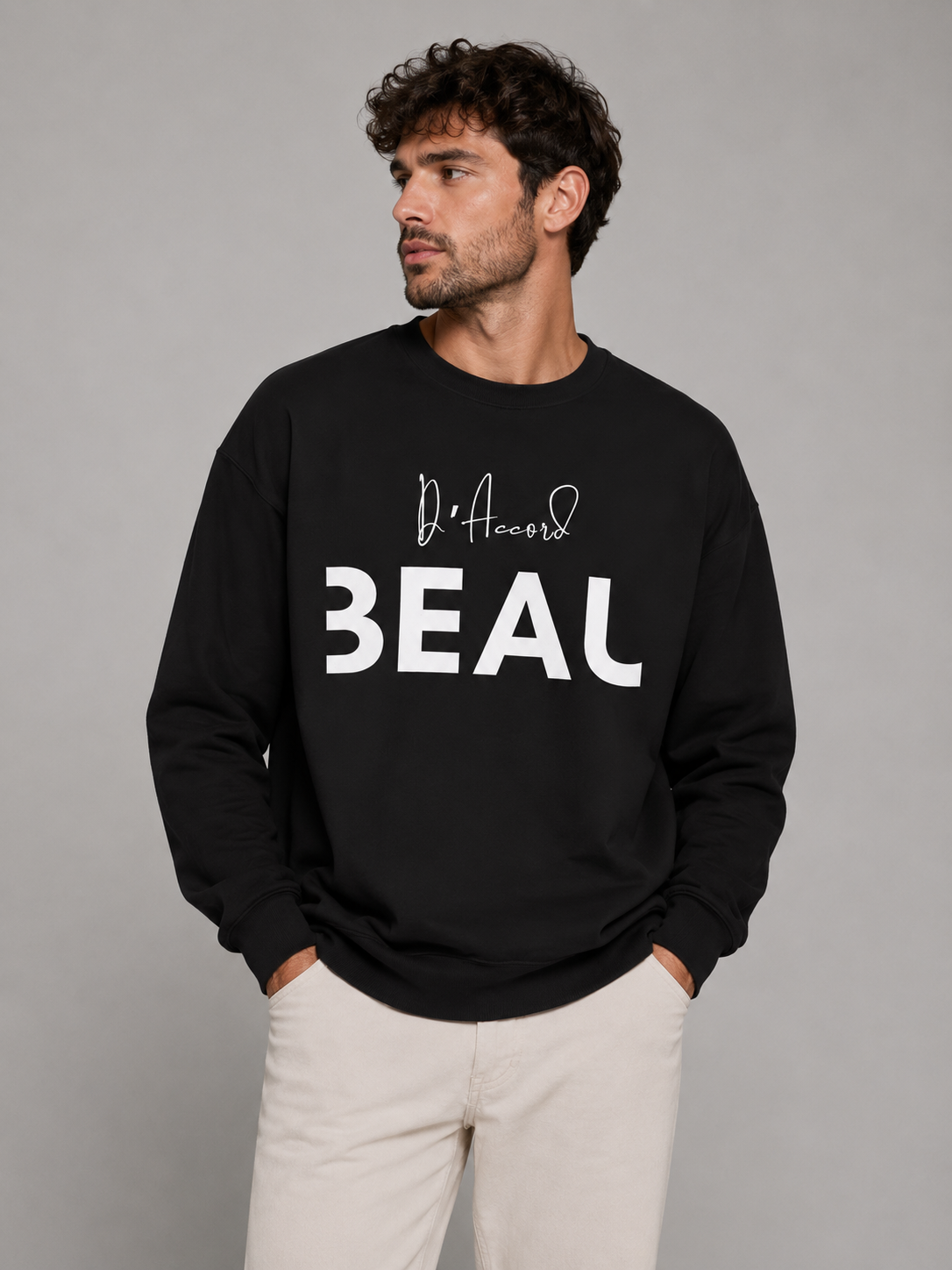 A man wearing a black crewneck sweatshirt featuring a black “D’accord” script above bold “BEAU” lettering across the chest, paired with beige trousers, against a light gray studio background.