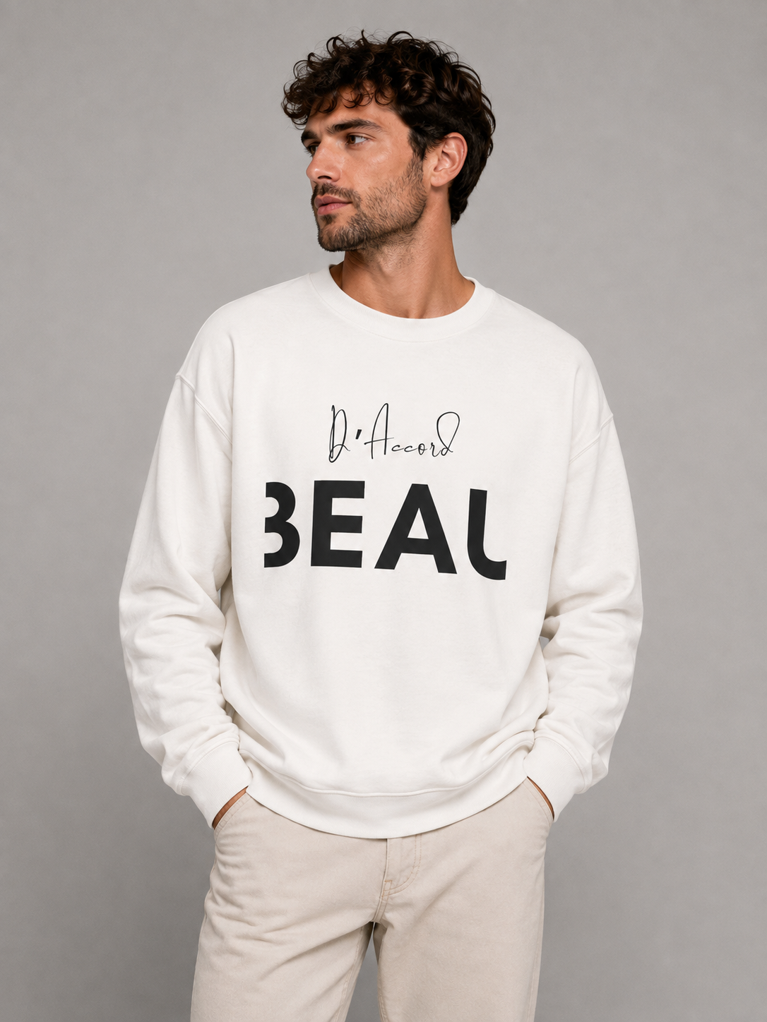 A man wearing a white crewneck sweatshirt featuring a black “D’accord” script above bold “BEAU” lettering across the chest, paired with beige trousers, against a light gray studio background.