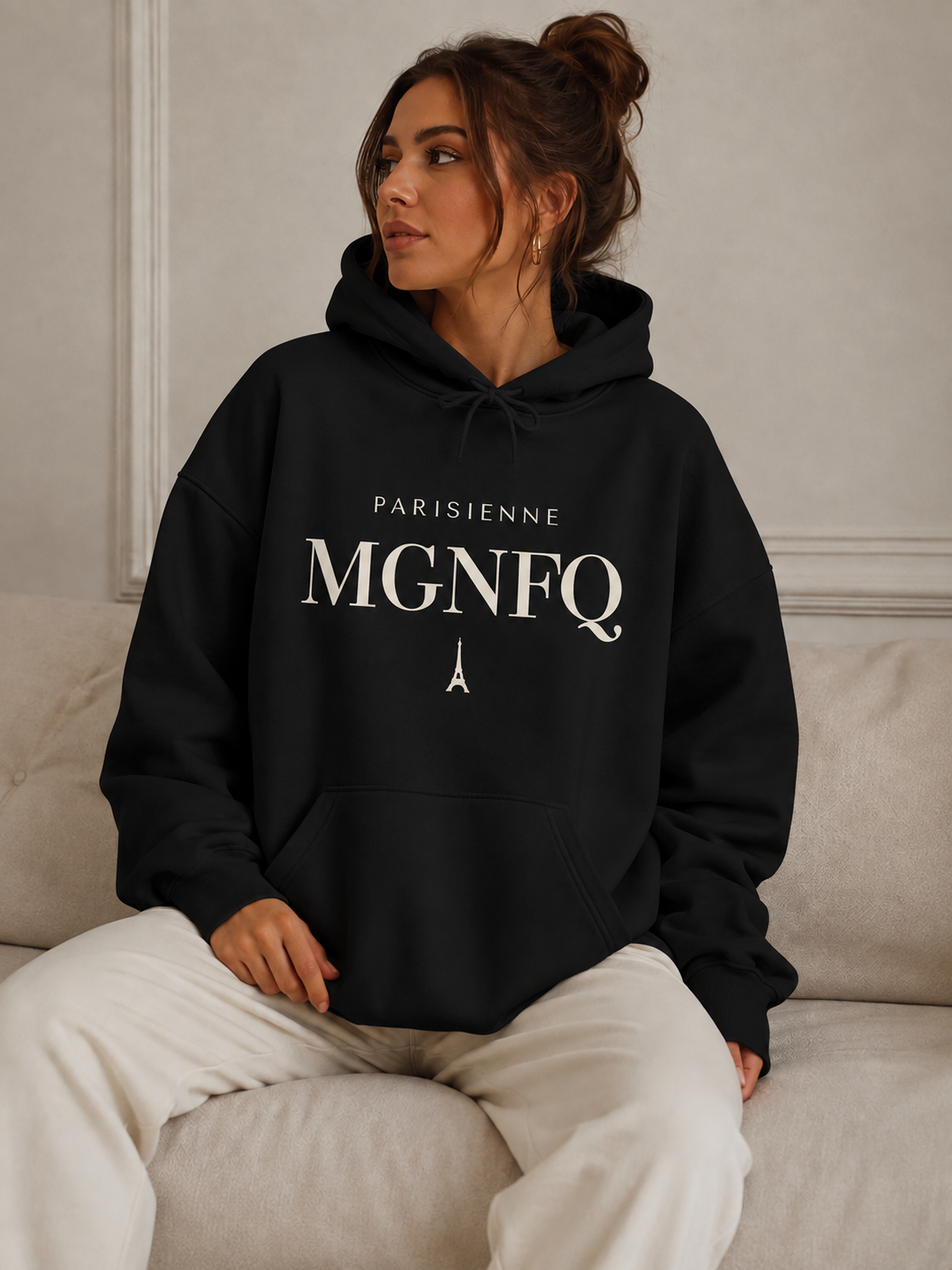 Woman wearing a black PARISIENNE MGNFQ graphic hoodie with cream typography and Eiffel Tower detail, styled with light trousers in a neutral editorial setting.