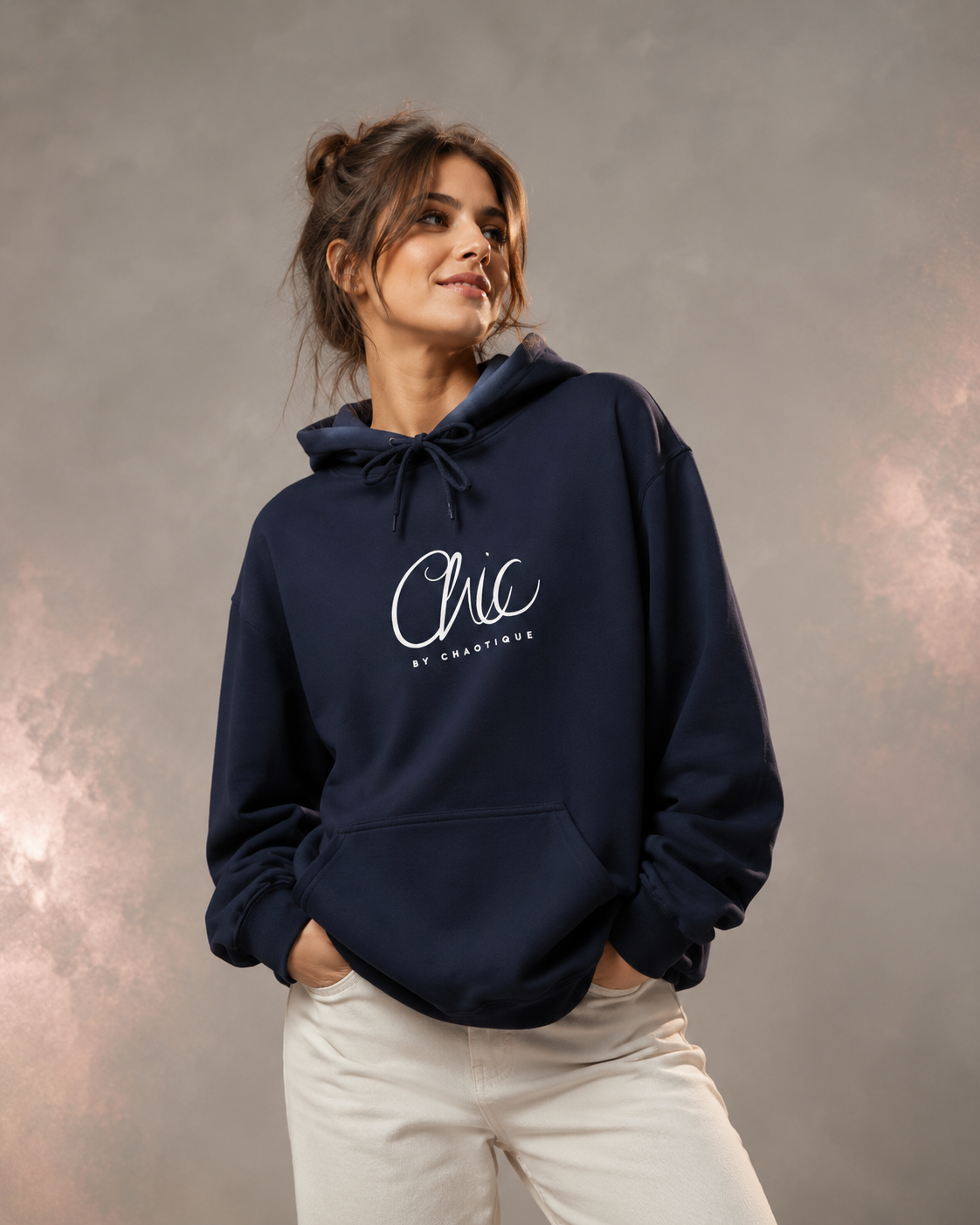 A woman wearing a navy blue Chic by Chaotique pullover hoodie with an elegant white script graphic on the chest, paired with light beige trousers, posing against a soft gray studio background with subtle rose-gold accents.
