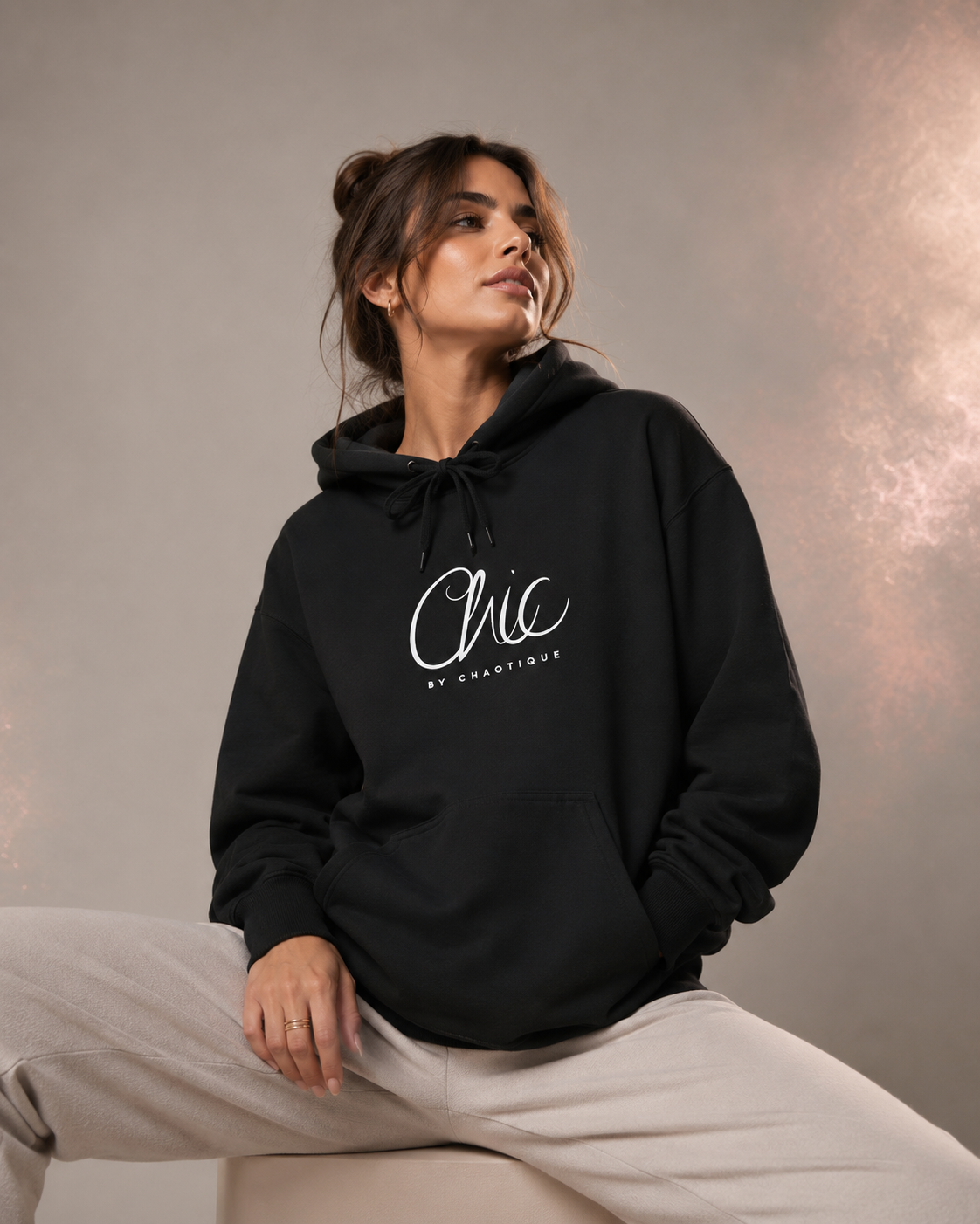 A woman wearing a black Chic by Chaotique pullover hoodie with an elegant white script graphic on the chest, paired with light beige trousers, posing against a soft gray studio background with subtle rose-gold accents.