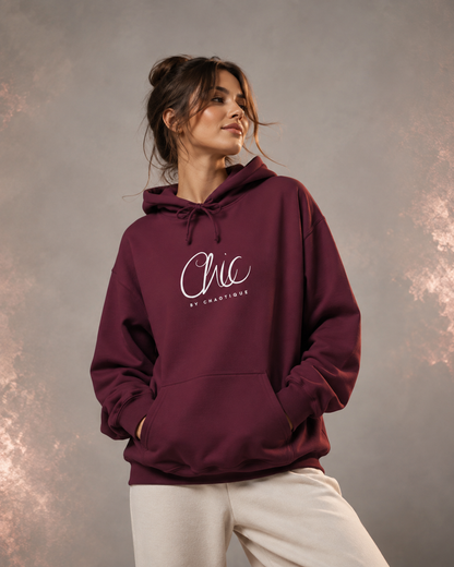A woman wearing a maroon Chic by Chaotique pullover hoodie with an elegant white script graphic on the chest, paired with light beige trousers, posing against a soft gray studio background with subtle rose-gold accents.