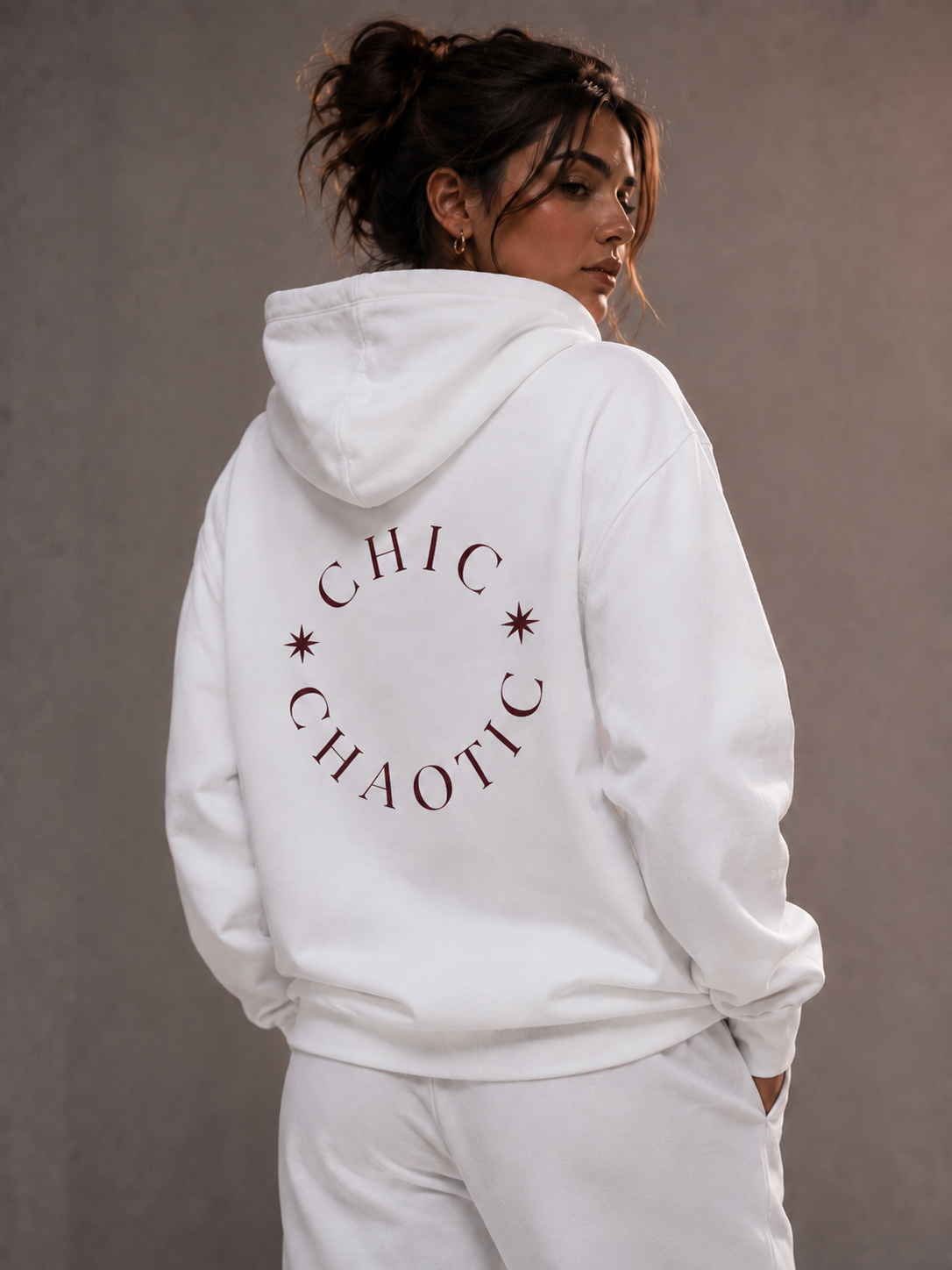 A woman wearing a white CHIC CHAOTIC graphic hoodie with dark red typography on the back, photographed from behind against a neutral gray studio background.