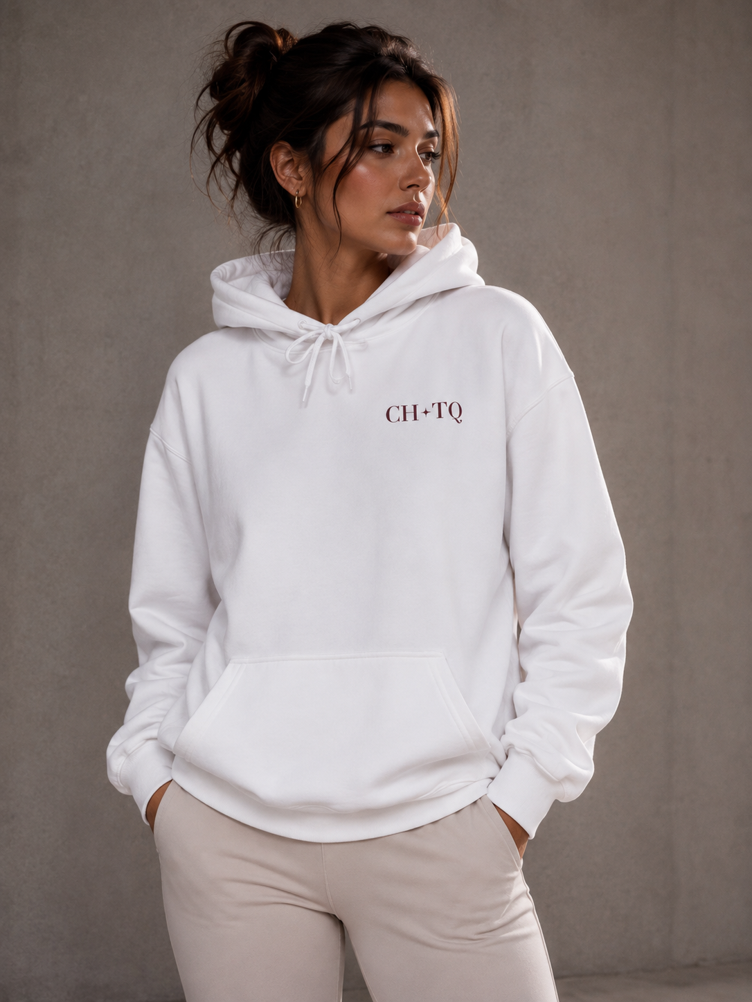 A woman wearing a white CH*TQ graphic hoodie with dark red typography, photographed in a neutral gray studio background.