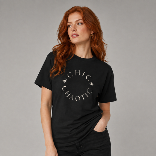 A woman with red hair wearing a black crewneck t-shirt with 'CHIC CHAOTIC' printed in white arched text with star accents on the chest, posed against a solid gray studio background.