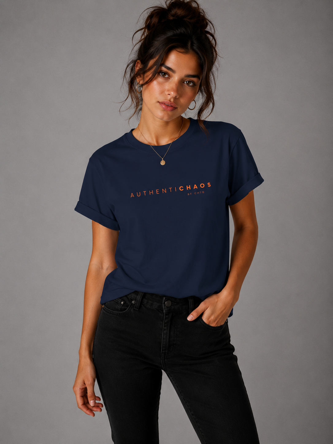 Woman wearing a navy blue AuthentiCHAOS by CHTQ graphic T-shirt with orange typography, paired with black jeans against a gray studio background.