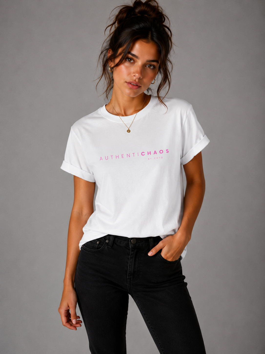 Woman wearing a white AuthentiCHAOS by CHTQ graphic T-shirt with pink typography, paired with black jeans against a gray studio background.
