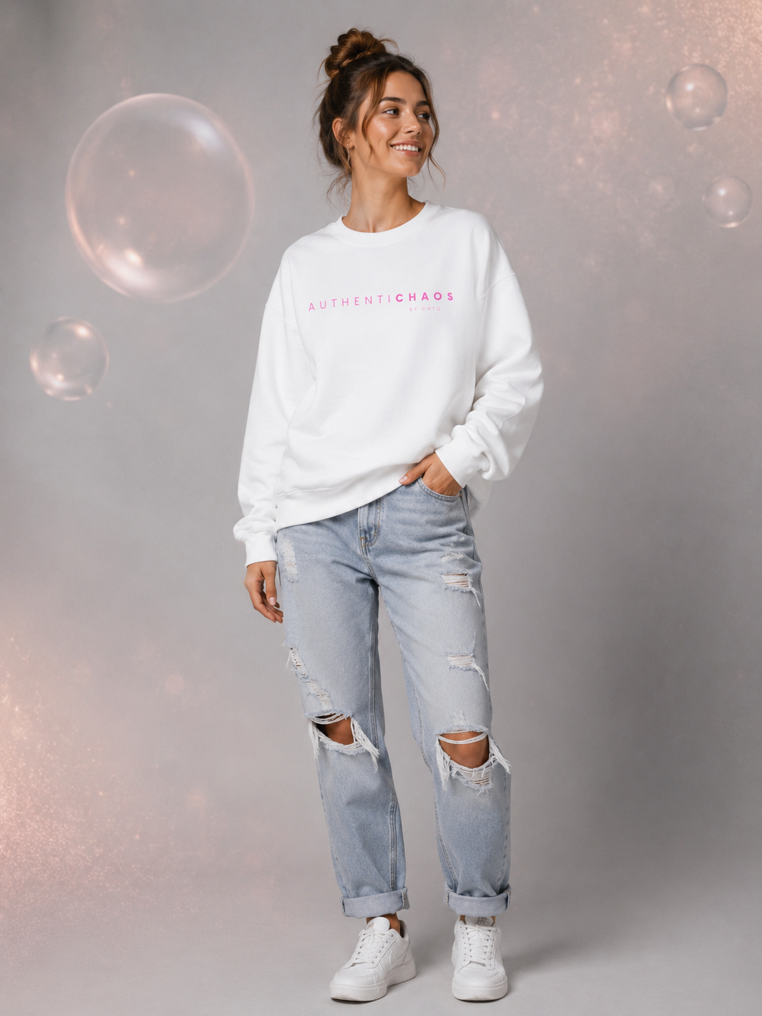 Woman wearing a white AuthentiCHAOS graphic sweatshirt with pink lettering in a modern gray studio setting.
