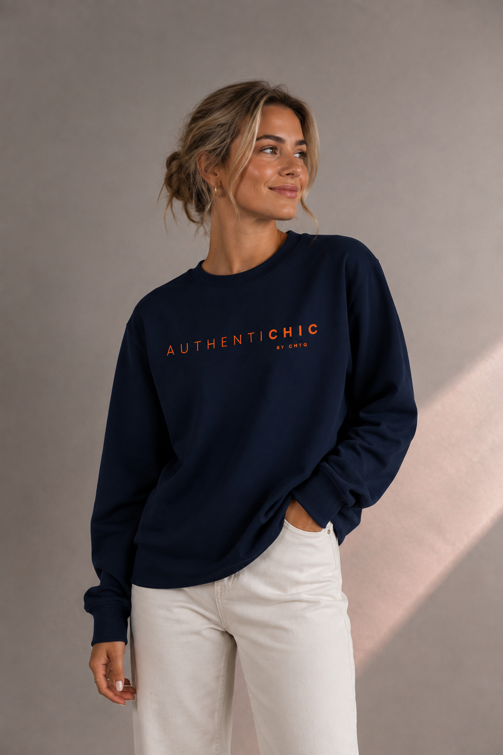 Young woman wearing a navy blue AuthentiCHIC graphic sweatshirt with orange lettering, styled with white trousers in a soft gray studio setting.