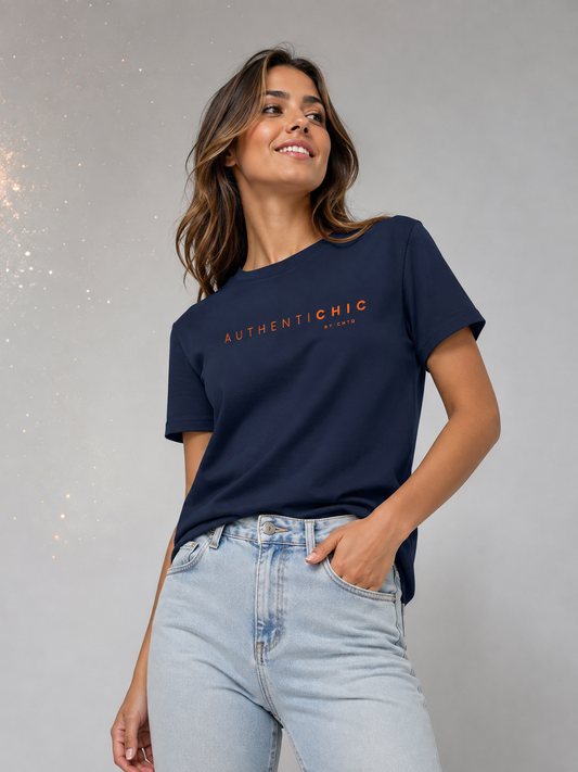 Young woman wearing a navy blue AuthentiCHIC graphic T-shirt with orange typography and light-wash jeans in a light gray studio with subtle golden sparkle details.
