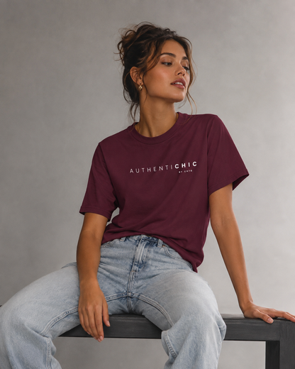 Young woman wearing a maroon AuthentiCHIC graphic T-shirt with orange typography and light-wash jeans in a light gray studio details.