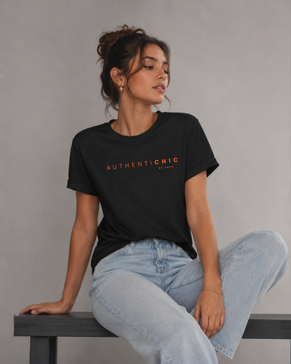 Young woman wearing a black AuthentiCHIC graphic T-shirt with orange typography and light-wash jeans in a light gray studio