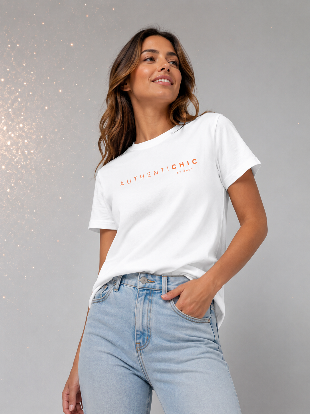 Young woman wearing a white AuthentiCHIC graphic T-shirt with orange typography and light-wash jeans in a light gray studio with subtle golden sparkle details.